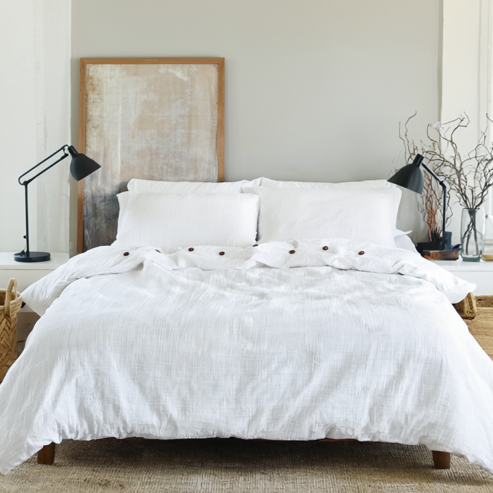 White bedroom with a neatly made bed with white organic duvet cover, lamps, and baskets.